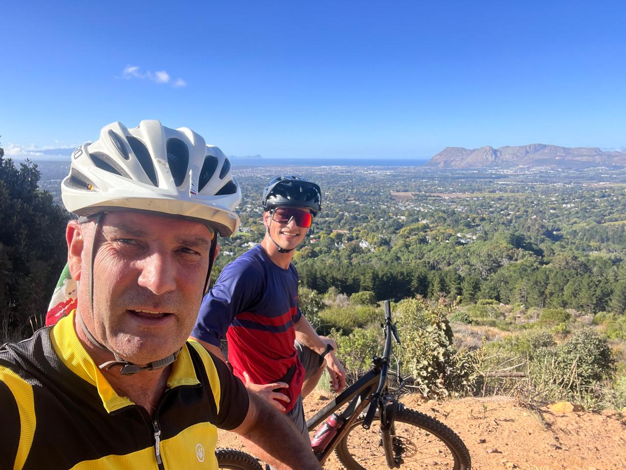 Two riders smiling for a selfie above a sweeping valley view under a blue sky